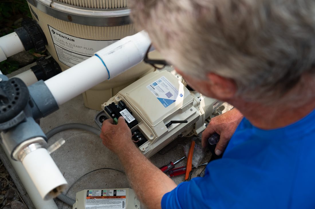 Technician in a blue shirt repairs a beige pool pump and filter system outdoors, wiring connections visible.